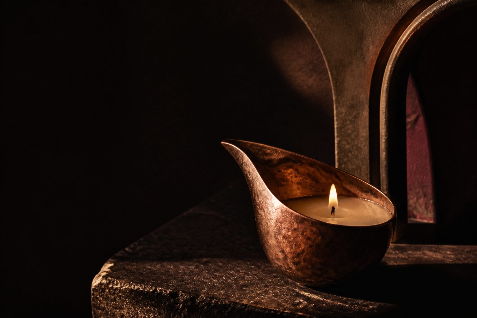 A candle burning in a sculptural copper vessel on dark stone.
